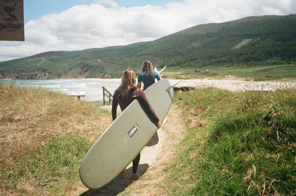 Two surfers walking along a dirt path toward the ocean with hills in the background, one carrying a light green O'Neill surfboard.