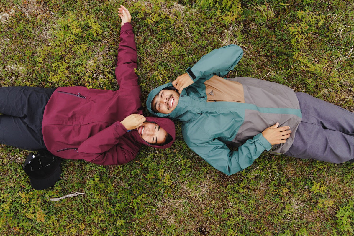 Two people lying down on the grass wearing waterproof jackets, hoods up, laughing