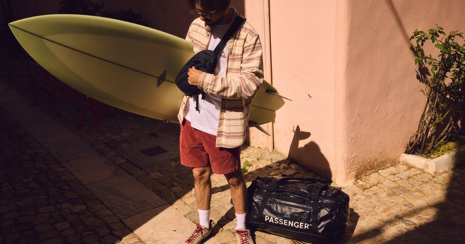 A man holds a surfboard under one arm, undoing his hip pack with the other and a duffel bag sits on the floor to his side